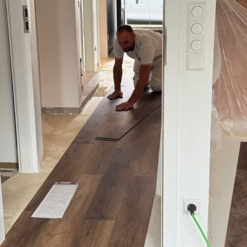 Ein Mann mit ernstem Blick verlegt braune Holzbodenplatten in einem Flur