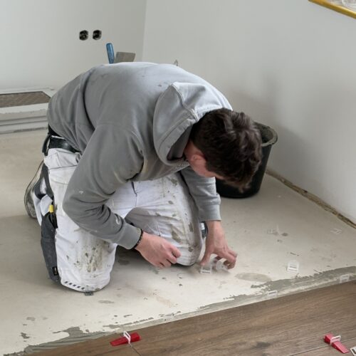 Ein Mann mit dunklem Haar kniet konzentriert beim Verlegen heller Fliesen am Boden