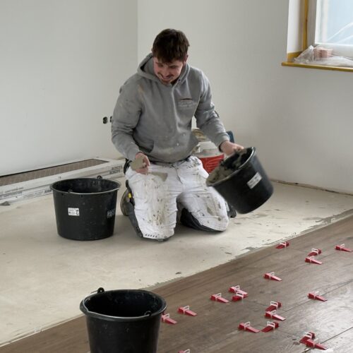 Ein junger Mann mit grauem Hoodie und weißen Arbeitshosen kniet lachend beim Verlegen von dunklem Laminat und hält einen schwarzen Eimer