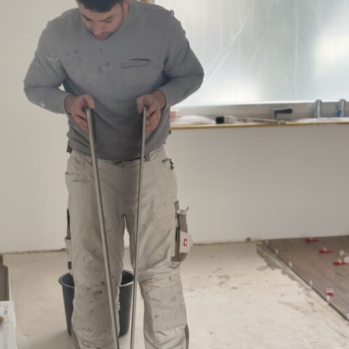 Ein Mann mit ernstem Blick trägt graues Shirt und weiße Hose in einem hellen Raum