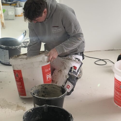 Ein junger Mann in grauem Hoodie kniet konzentriert vor mehreren Farbeimern im hellen Raum