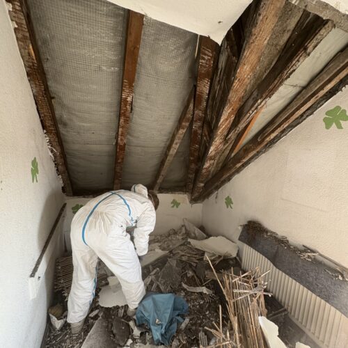 Eine Person im weißen Schutzanzug arbeitet konzentriert in einem beschädigten Raum