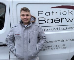 Junger Mann mit kurzem Haar und hellem Mantel steht freundlich vor einem weißen Firmenwagen