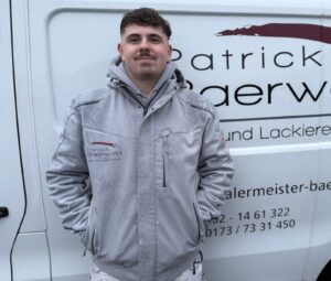 Junger Mann mit leichtem Lächeln trägt eine graue Jacke vor einem Firmenwagen