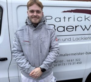 Ein junger Mann mit blondem Haar lächelt freundlich vor einem weißen Firmenwagen