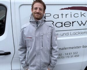 Ein lächelnder Mann mit roter Kurzhaarfrisur steht vor einem weißen Firmenwagen