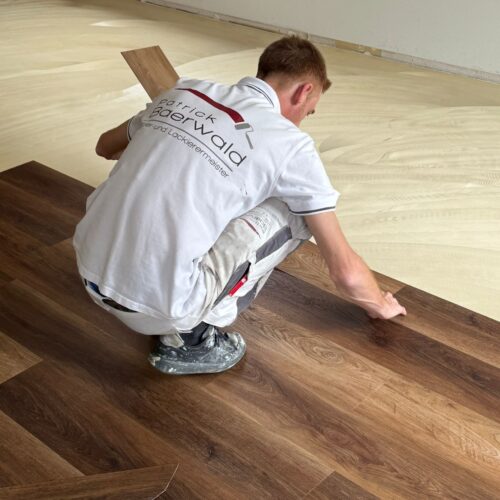 Ein junger Mann kniet konzentriert auf hellem Boden und verlegt dunkles Parkett