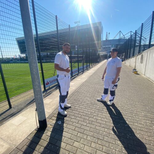 Zwei Männer in weißer Arbeitskleidung stehen lachend vor einem modernen Stadion im Sonnenschein